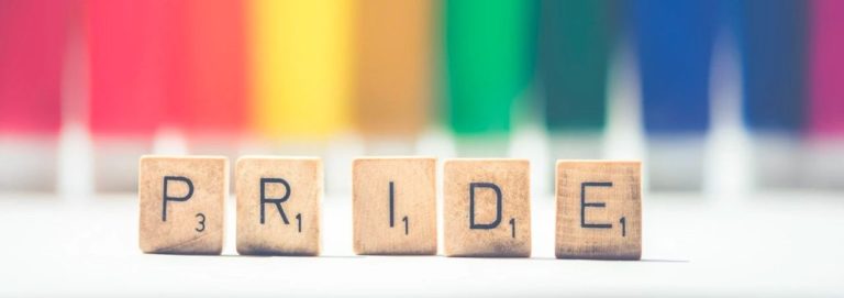 Scrabble-like tiles spelling “PRIDE” in front of a rainbow flag.