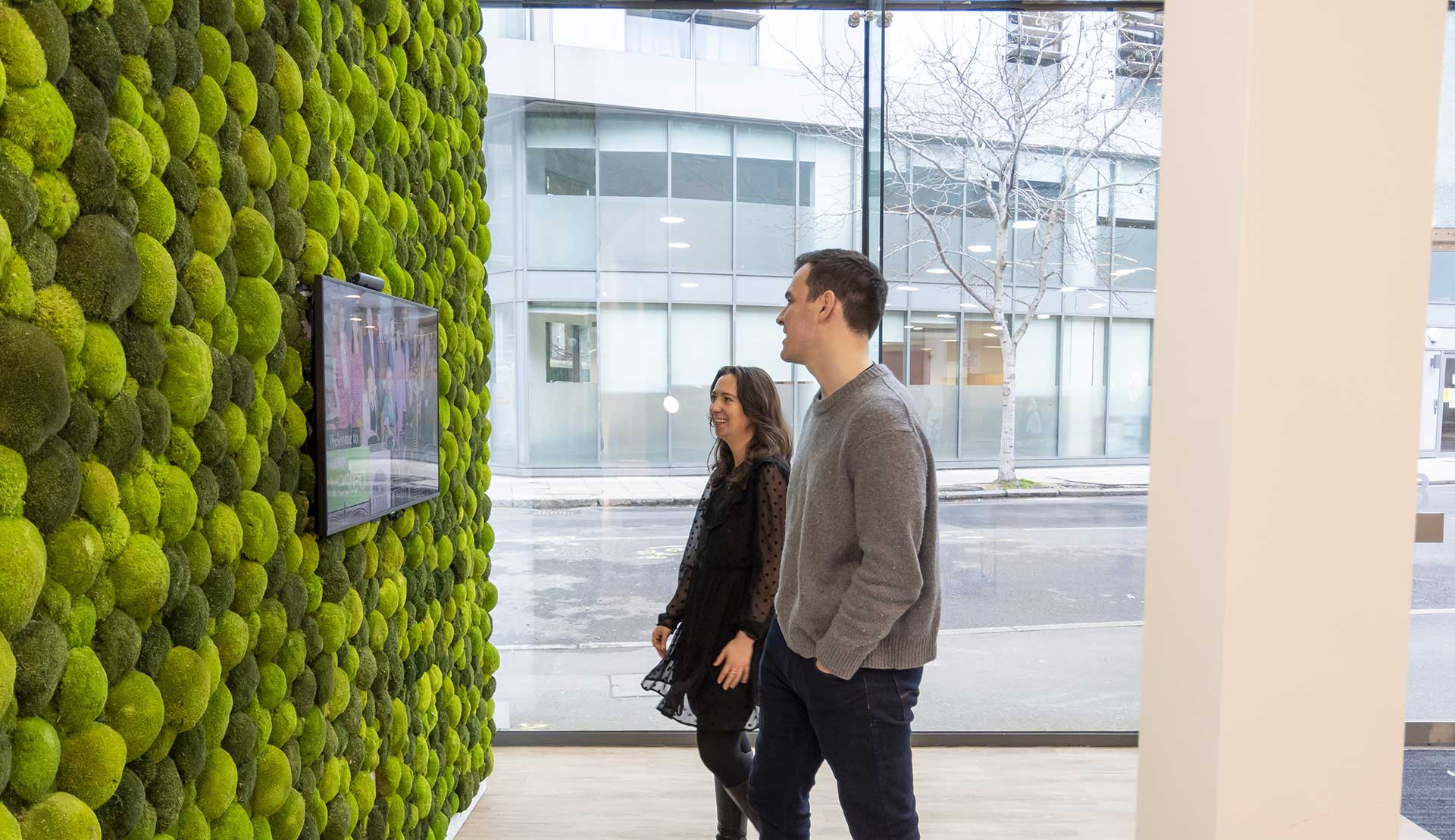 A man and a woman in the entry way of the London headquarters viewing a monitor screen mounted on a textured green wall.