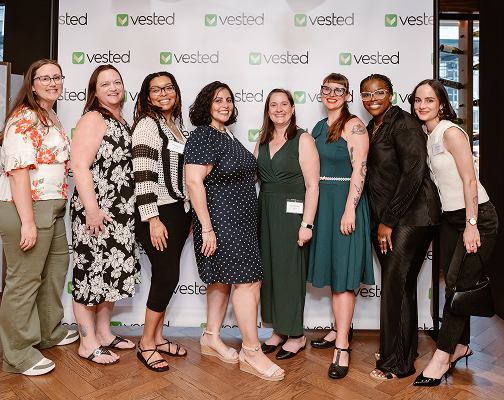 Eight Vested team members smiling together in front of a Vested branded backdrop at a company event